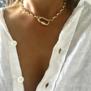 Pu&ograve; includere: Una collana a catena color oro con una grande chiusura. La collana presenta un design a maglie grosse e una chiusura scintillante ricoperta di cristalli. La collana &egrave; indossata sopra una camicia bianca abbottonata.
