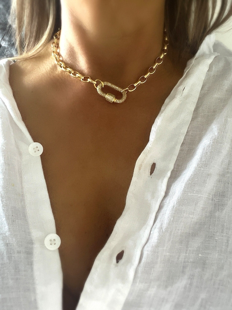 Pu&ograve; includere: Una collana a catena color oro con una grande chiusura. La collana presenta un design a maglie grosse e una chiusura scintillante ricoperta di cristalli. La collana &egrave; indossata sopra una camicia bianca abbottonata.