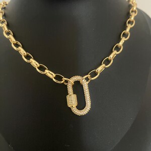 Pu&ograve; includere: Una collana color oro con un grande pendente di forma ovale. Il pendente e le maglie della catena sono collegati. Il pendente &egrave; incrostato di piccole pietre trasparenti. La collana &egrave; esposta su una superficie nera.