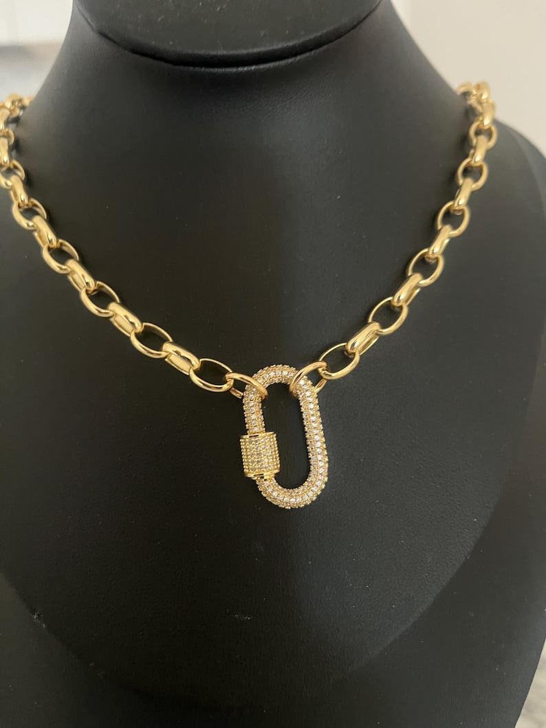 Pu&ograve; includere: Una collana color oro con un grande pendente di forma ovale. Il pendente e le maglie della catena sono collegati. Il pendente &egrave; incrostato di piccole pietre trasparenti. La collana &egrave; esposta su una superficie nera.