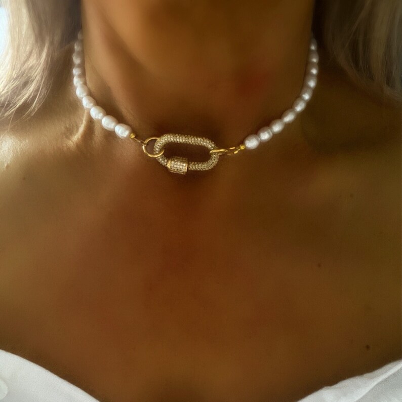 May include: A white pearl necklace with a gold clasp featuring a row of small clear stones. The necklace is worn by a person.