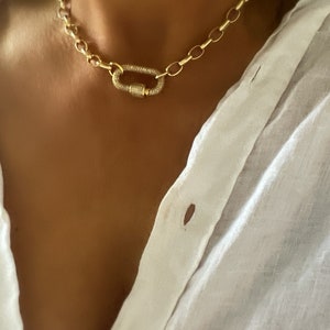 Pu&ograve; includere: Una collana a catena dorata con una grande chiusura ovale. La chiusura &egrave; incastonata con piccole pietre preziose trasparenti. La collana &egrave; indossata su una camicia bianca.