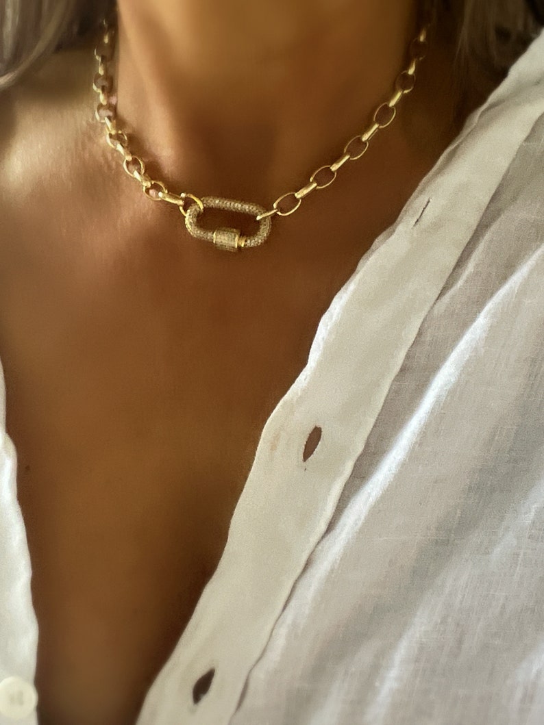 Pu&ograve; includere: Una collana a catena dorata con una grande chiusura ovale. La chiusura &egrave; incastonata con piccole pietre preziose trasparenti. La collana &egrave; indossata su una camicia bianca.
