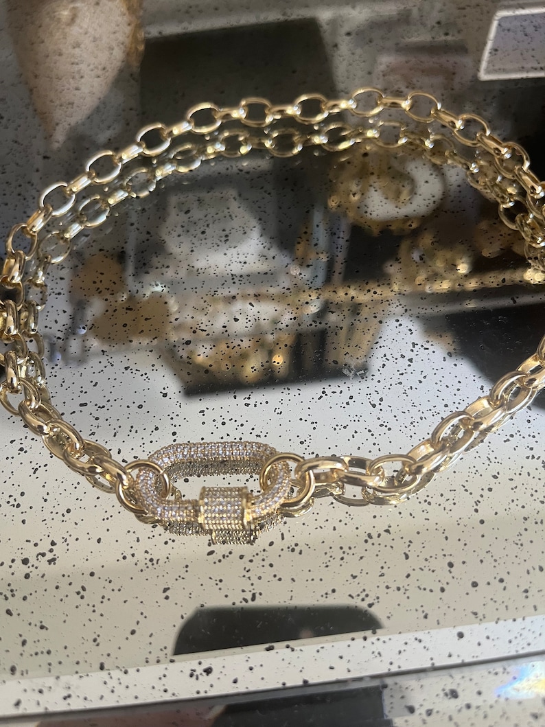 Pu&ograve; includere: Collana a catena color oro con una grande chiusura decorata. La collana presenta un design a catena a doppio filo con maglie ovali. La chiusura &egrave; incastonata con piccoli cristalli trasparenti, che aggiungono brillantezza al gioiello.