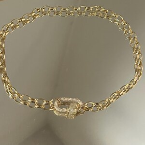 Pu&ograve; includere: Collana a catena dorata con chiusura decorativa. La collana presenta un design a doppia catena con maglie ovali. La chiusura &egrave; di forma rettangolare, incastonata con piccoli cristalli trasparenti. La collana &egrave; esposta su una superficie riflettente.