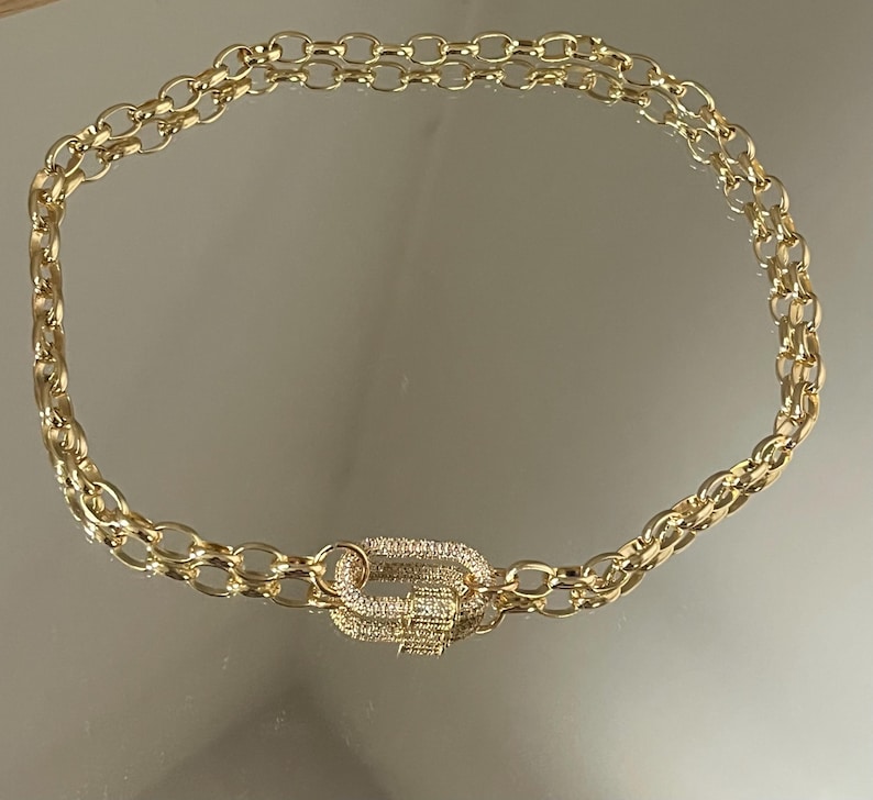 Pu&ograve; includere: Collana a catena dorata con chiusura decorativa. La collana presenta un design a doppia catena con maglie ovali. La chiusura &egrave; di forma rettangolare, incastonata con piccoli cristalli trasparenti. La collana &egrave; esposta su una superficie riflettente.