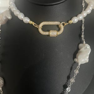 May include: A layered necklace with a large, oval-shaped pendant featuring a pave setting of clear rhinestones. The necklace is made of white pearls and silver chain with clear rhinestones.