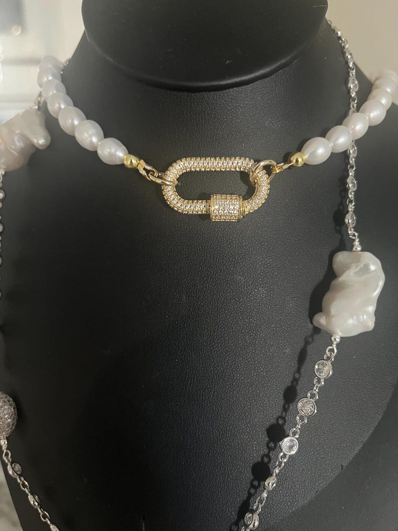 May include: A layered necklace with a large, oval-shaped pendant featuring a pave setting of clear rhinestones. The necklace is made of white pearls and silver chain with clear rhinestones.