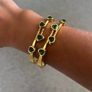 May include: Three gold bangles with green heart-shaped stones. The bangles are stacked on a person's wrist.