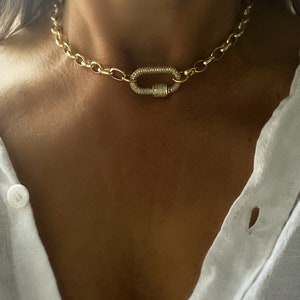 Pu&ograve; includere: Collana a catena color oro con una grande chiusura ovale. La chiusura &egrave; incastonata con piccoli cristalli scintillanti. La collana ha un design a maglie grosse e si indossa intorno al collo.