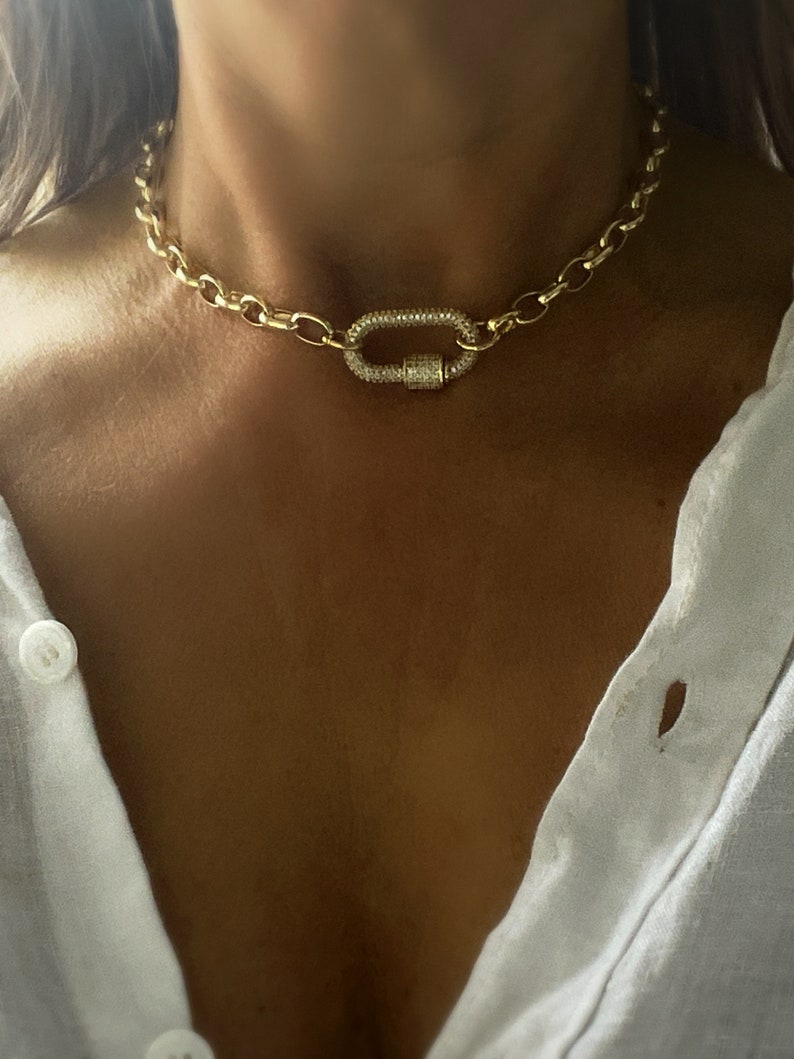 Pu&ograve; includere: Collana a catena color oro con una grande chiusura ovale. La chiusura &egrave; incastonata con piccoli cristalli scintillanti. La collana ha un design a maglie grosse e si indossa intorno al collo.