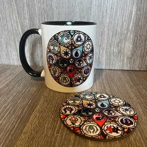 May include: A white ceramic mug with a black rim and handle features a colourful stained glass design of Magic: The Gathering symbols. The mug is paired with a matching coaster.