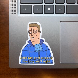 May include: A sticker featuring a cartoon character with a blue jacket and a scarf, with the text "6:00 AM and already the boy ain't right".