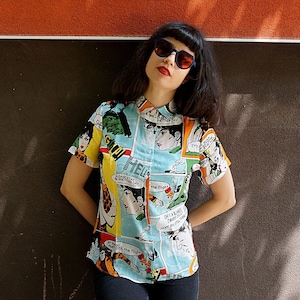 May include: A woman wearing a blue, short-sleeved shirt with a comic book print. The shirt features colorful illustrations and text, including "Please stay another day" and "It's the flight". She is wearing black sunglasses and has dark hair.