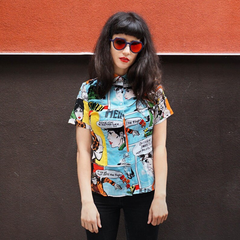 May include: A woman wearing a blue, short-sleeved button-down shirt with a comic book print. The shirt features a variety of colorful characters and text, including "Please stay for another day" and "Oh I'm always thinking how this still works". She is also wearing black pants and sunglasses.