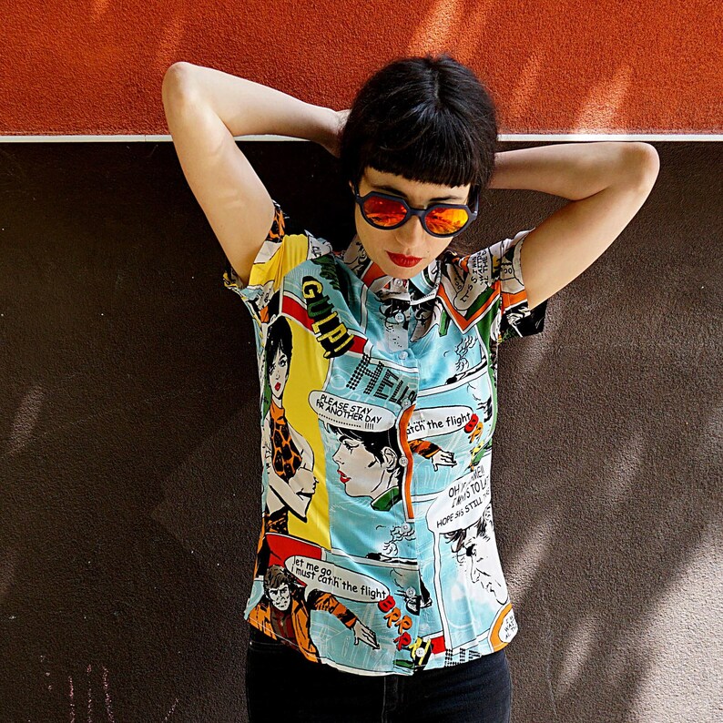 May include: A blue and yellow short-sleeved shirt with a comic book print. The shirt features a variety of characters and text, including "Gulpi", "Please stay for another day", "Catch the flight", and "Oh, it's too late, hope we still make it".
