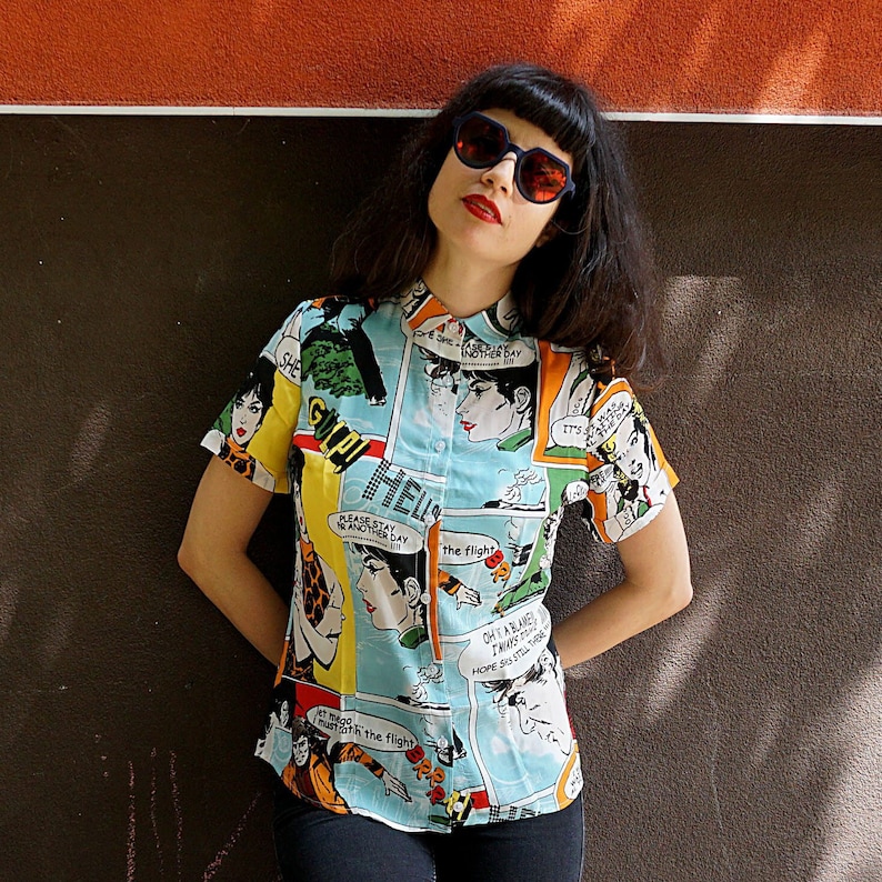 May include: A woman wearing a blue, short-sleeved shirt with a comic book print. The shirt features colorful illustrations and text, including "Please stay another day" and "It's the flight". She is wearing black sunglasses and has dark hair.