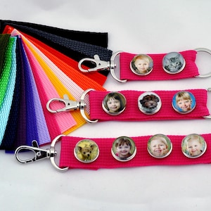 Könnte beinhalten: Drei rosa Schlüsselanhänger mit silbernen Schlüsselringen und Fotoeinsätzen. Jeder Schlüsselanhänger hat ein anderes Foto einer Person oder eines Tieres. Die Schlüsselanhänger werden auf einer weißen Oberfläche mit einer Vielzahl von farbigen Stoffstreifen präsentiert.