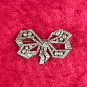May include: A silver bow brooch with clear rhinestones set in a geometric pattern. The brooch is displayed against a red velvet backdrop, emphasising its intricate details and vintage style.
