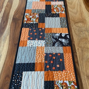 May include: A black and orange Halloween table runner with a patchwork design featuring pumpkins, ghosts, and polka dots. The runner has a black border.