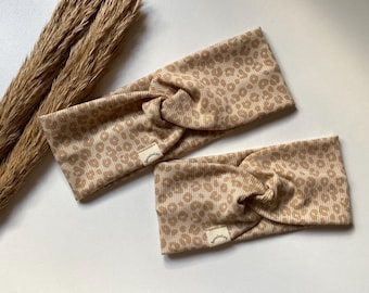 May include: Two beige headbands with a leopard print pattern. The headbands have a twisted knot design and small rectangular tags. Dried wheat stalks are in the background.