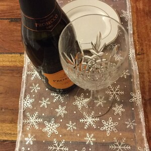 Può includere: Una bottiglia di prosecco con un runner da tavolo con motivo a fiocco di neve bianco e un bicchiere trasparente.