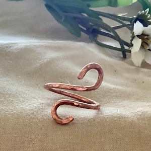 Könnte beinhalten: Handgefertigter Kupferring mit gehammerter Textur. Der Ring hat ein einzigartiges, offenes Design mit zwei gebogenen Enden, wodurch eine Spiralform entsteht. Der Ring wird auf einem neutralfarbenen Stoff mit Grün und weißen Blüten im Hintergrund präsentiert.