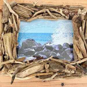 May include: A square picture frame made from natural driftwood. The frame surrounds a photo of a blue ocean scene with waves crashing against dark rocks. The driftwood is various shades of brown and beige.