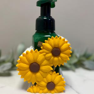May include: A green bottle with a pump dispenser, adorned with three yellow sunflowers with brown centers. The sunflowers are clustered together, creating a decorative accent. The bottle is likely for liquid soap or lotion.