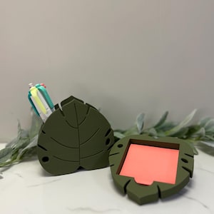 Puede incluir: Organizador de escritorio con forma de hoja verde oliva con interior color coral. El organizador está abierto, revelando el interior, y otro está en posición vertical, sosteniendo bolígrafos. El diseño presenta vetas de hojas y un detalle recortado.