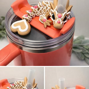 May include: A coral travel mug with a decorative dragon lid. The dragon is tan with white and pink details, and a clear straw protrudes from the lid. A heart-shaped biscuit is also on the lid.