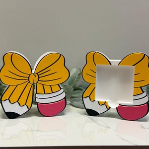 Puede incluir: Dos accesorios de lazo con forma de lápiz. Cada uno tiene un lazo amarillo, contornos negros, una goma de borrar blanca y una punta de lápiz rosa. Uno tiene un cuadrado blanco en el centro. Los accesorios están sobre una superficie blanca.