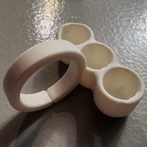 Może przedstawiać: Biały, drukowany w 3D pierścień i obiekt z wieloma otworami. Pierścień to solidna opaska z otworem na zawiasach. Drugi obiekt ma trzy okrągłe wgłębienia. Oba przedmioty wykonane są z matowego, kremowego materiału.