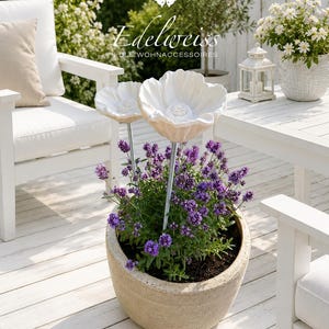 Könnte beinhalten: Ein runder, hellbeiger Steintopf mit einer leuchtend violetten Lavendelpflanze und zwei dekorativen weißen Blumensteckern aus Keramik. Die Stecker haben große, geformte weiße Blütenblätter mit strukturierten Mitten auf dünnen, silberfarbenen Metallstäben.