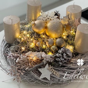 Könnte beinhalten: Ein festlicher Weihnachtskranz mit vier goldenen Kerzen, dekorativen Ornamenten und Lichterketten. Der Kranz besteht aus weißen Zweigen und ist mit Tannenzapfen, Beeren und einem Holzstern mit dem Wort "Xmas" verziert.
