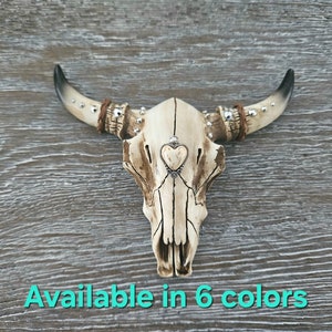 May include: A white ceramic bull skull with silver accents and a small heart charm hanging from the center. The skull is on a wood background. Available in 6 colors.