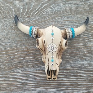 May include: A white ceramic bull skull with turquoise and silver accents. The skull has a decorative silver filigree design on the forehead and turquoise beads along the center line.