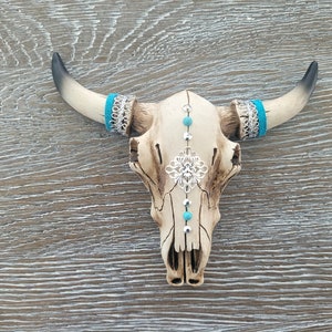 May include: A white ceramic bull skull with turquoise and silver accents. The skull has a decorative silver filigree design on the forehead and turquoise beads along the center line.