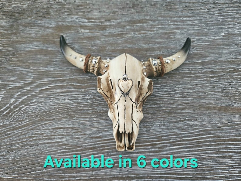 May include: A white ceramic bull skull with silver accents and a small heart charm hanging from the center. The skull is on a wood background. Available in 6 colors.