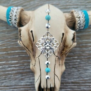 May include: A white ceramic bull skull with turquoise and silver accents. The skull has a silver filigree design in the center and turquoise beads and silver studs down the center.