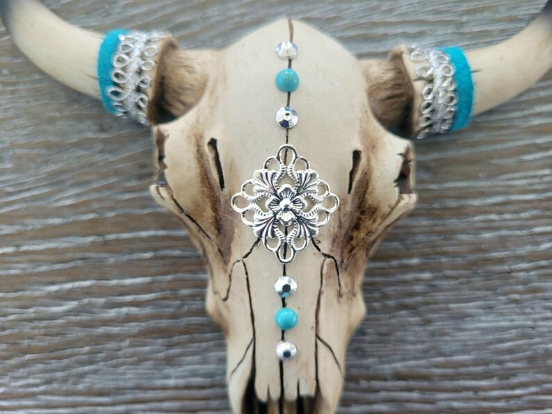 May include: A white ceramic bull skull with turquoise and silver accents. The skull has a silver filigree design in the center and turquoise beads and silver studs down the center.