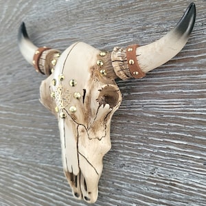 Peut inclure: Un cr&acirc;ne de taureau en c&eacute;ramique blanche avec des accents dor&eacute;s et des lani&egrave;res en cuir marron. Le cr&acirc;ne est mont&eacute; sur un mur en bois.