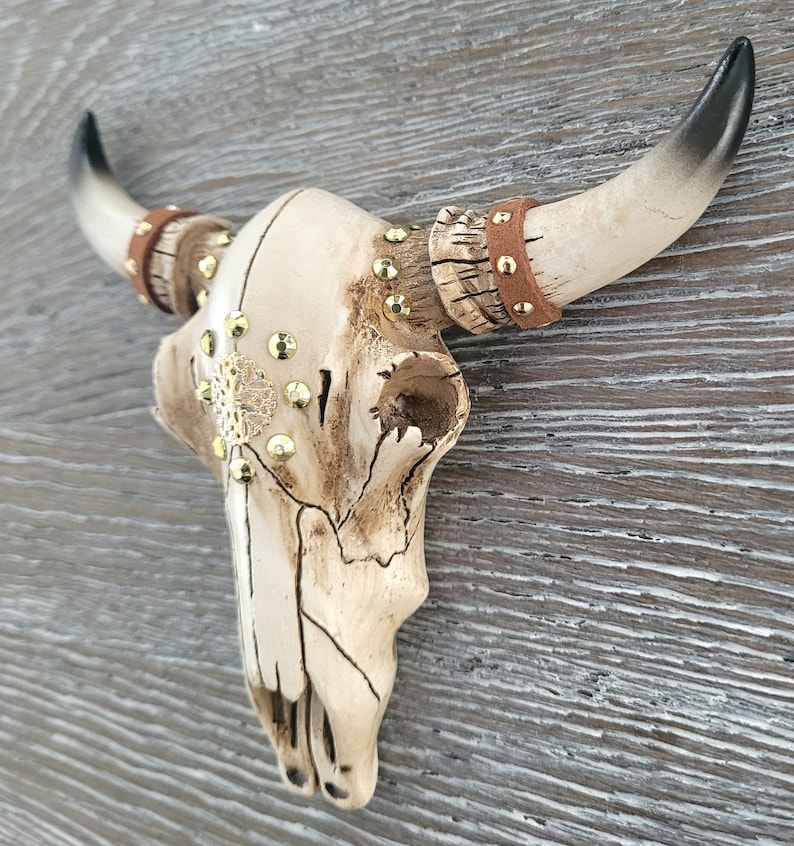 Peut inclure: Un cr&acirc;ne de taureau en c&eacute;ramique blanche avec des accents dor&eacute;s et des lani&egrave;res en cuir marron. Le cr&acirc;ne est mont&eacute; sur un mur en bois.