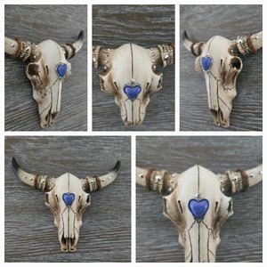 May include: A white ceramic bull skull with silver accents and blue heart-shaped gems on each side. The skull is mounted on a wooden wall.
