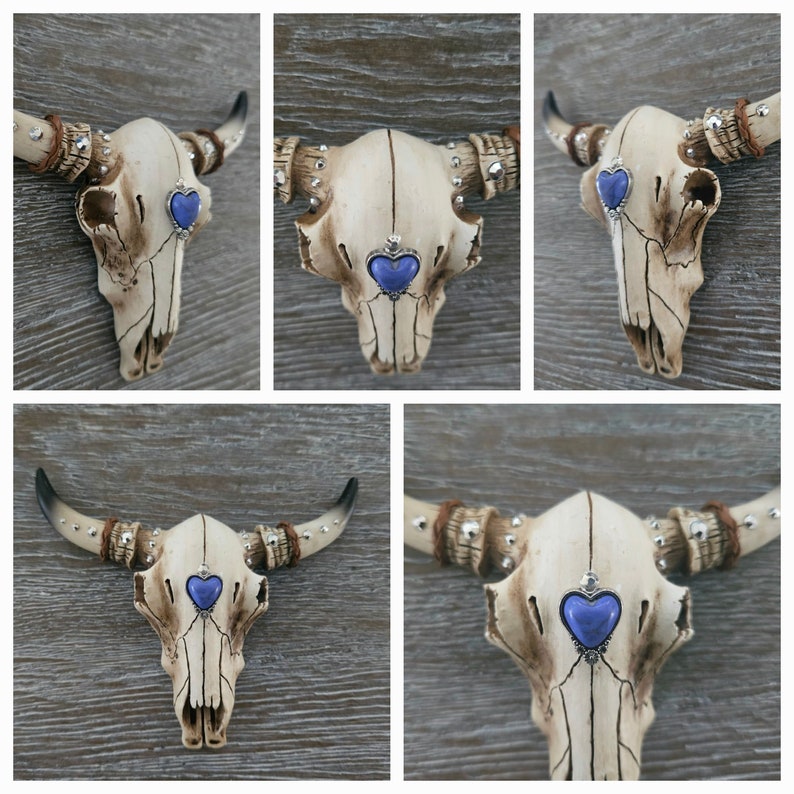 May include: A white ceramic bull skull with silver accents and blue heart-shaped gems on each side. The skull is mounted on a wooden wall.