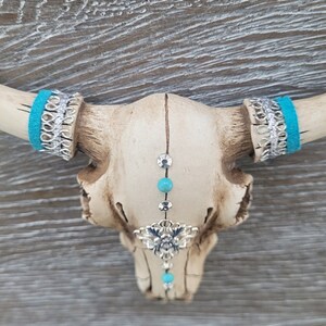 May include: A white ceramic bull skull with turquoise and silver accents. The skull has a beaded chain hanging from the center of the forehead with a silver charm.