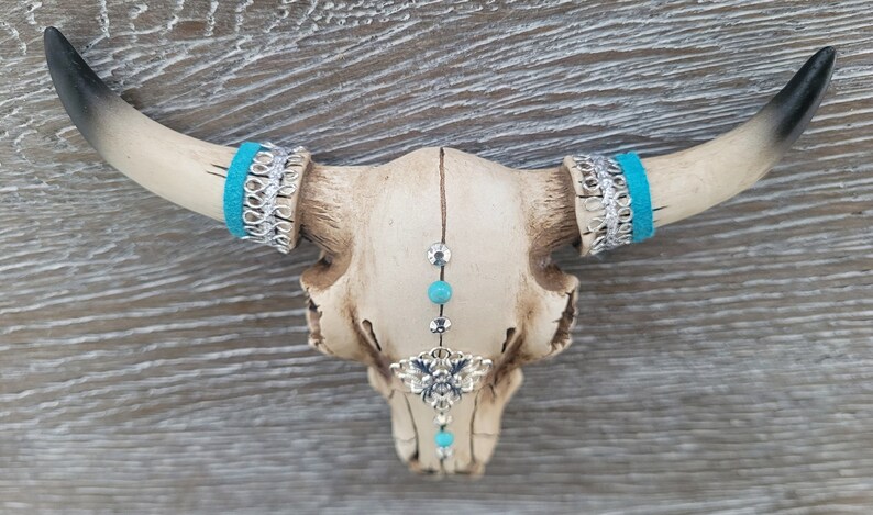 May include: A white ceramic bull skull with turquoise and silver accents. The skull has a beaded chain hanging from the center of the forehead with a silver charm.