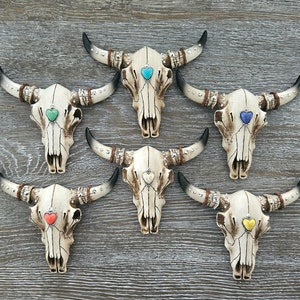 May include: Six white ceramic bull skulls with different colored heart accents. The skulls are decorated with silver studs and have horns.