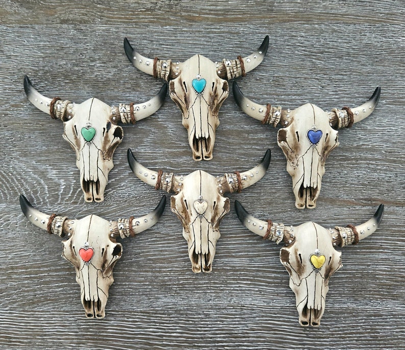 May include: Six white ceramic bull skulls with different colored heart accents. The skulls are decorated with silver studs and have horns.