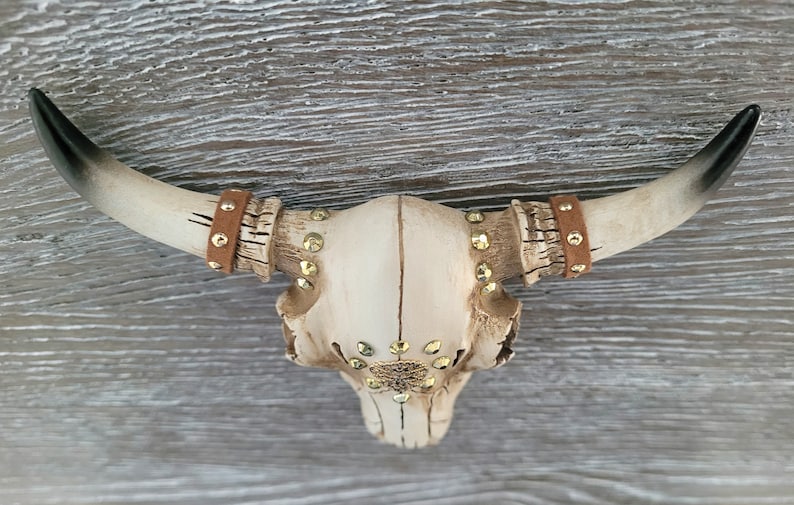 Peut inclure: Un cr&acirc;ne de taureau en c&eacute;ramique blanche avec des accents dor&eacute;s et des lani&egrave;res en cuir marron. Le cr&acirc;ne est d&eacute;cor&eacute; de strass et a un style rustique et western.